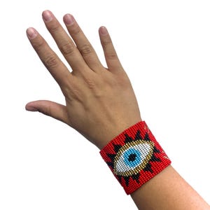 May include: A wide cuff bracelet made of small glass beads in a red, black, white, gold, and blue color palette. The beadwork features a central eye motif with a blue pupil, white iris, and gold outline, surrounded by black rays on a red background.