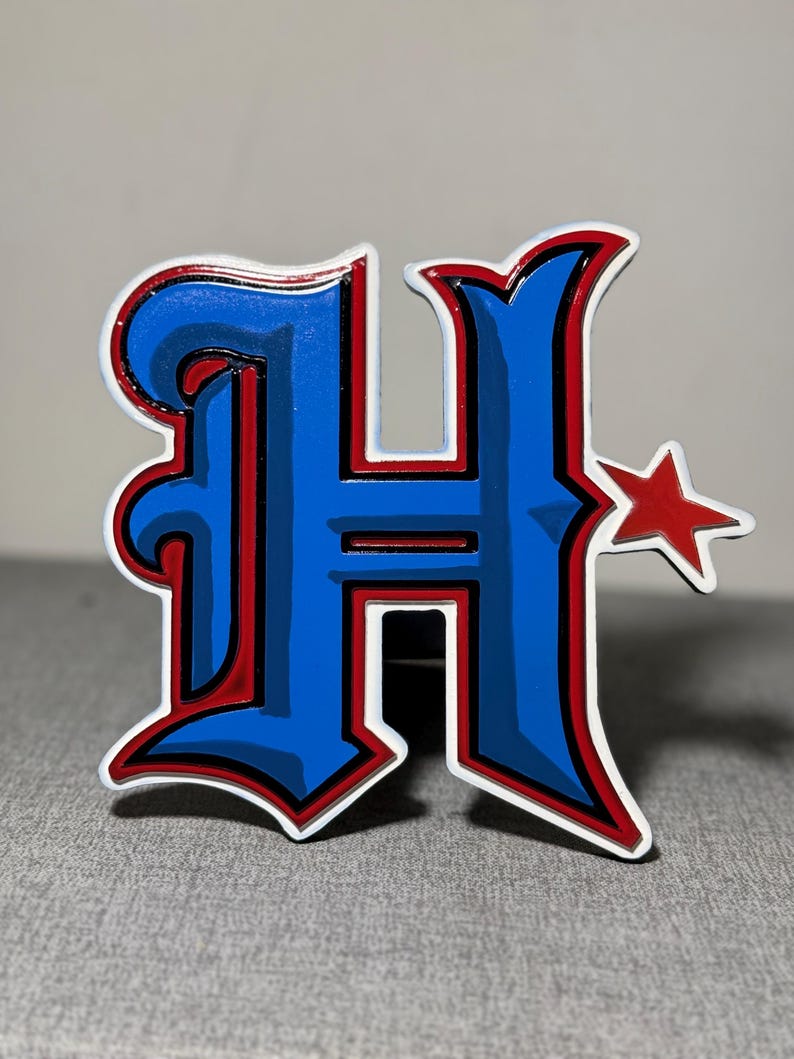May include: A blue and red enamel pin with a stylized letter "H" and a red star. The pin has a white outline.
