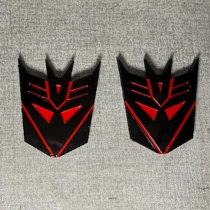 Puede incluir: Dos emblemas idénticos con el logotipo Decepticon en negro y rojo de la franquicia Transformers. Cada emblema tiene una forma de escudo angular y afilada con un diseño de cara central, orejas puntiagudas y recortes geométricos en rojo.
