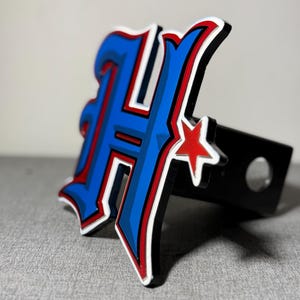 May include: A blue and red stylized letter H with a red star, attached to a black hitch cover.