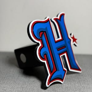 May include: A blue and red stylized letter "H" with a white outline and a small red star. The letter is attached to a black metal hitch cover.