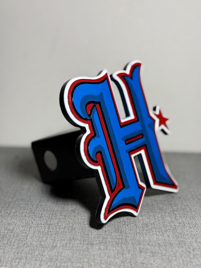 May include: A blue and red stylized letter "H" with a white outline and a small red star. The letter is attached to a black metal hitch cover.