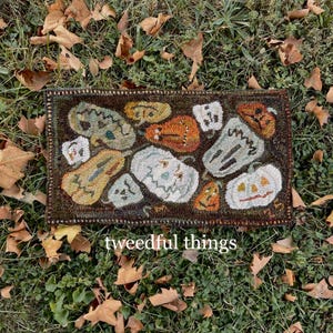 May include: A rectangular, hand-stitched rug featuring various Halloween-themed pumpkins in shades of orange, white, and green. The rug has a dark brown border and is set against a backdrop of grass and fallen leaves. The text "tweedful things" is visible.