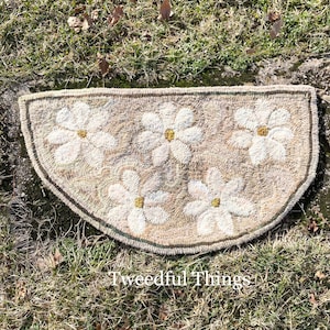 May include: A half-round rug with a beige background and a white floral pattern. The rug has a green border and is made of wool. The rug is on a bed of green grass.