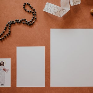 May include: A flat lay of religious items on a brown surface. There is a rosary, a wooden cross, two white pieces of paper, and a white ribbon with a repeating pattern of a dove.