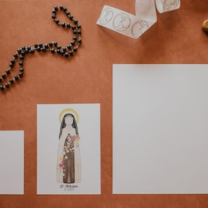 Puede incluir: Una tarjeta blanca con una ilustraci&oacute;n de acuarela de Santa Teresa de Lisieux, una santa cat&oacute;lica, con el texto "St. Therese of Lisieux" y "Little Flower". La tarjeta est&aacute; sobre una superficie marr&oacute;n con un rosario, una cinta blanca con c&iacute;rculos y un crucifijo de madera.