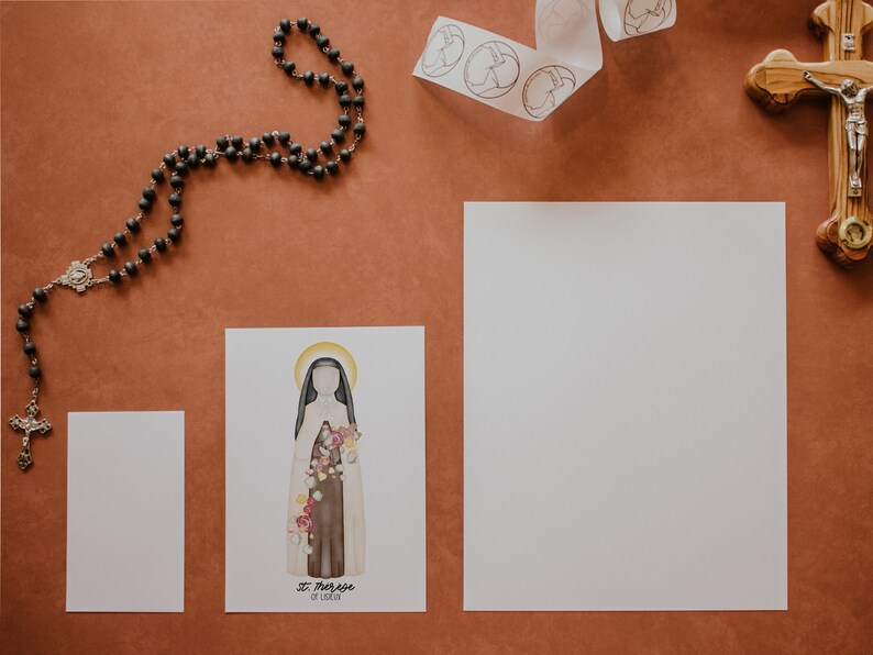 Puede incluir: Una tarjeta blanca con una ilustraci&oacute;n de acuarela de Santa Teresa de Lisieux, una santa cat&oacute;lica, con el texto "St. Therese of Lisieux" y "Little Flower". La tarjeta est&aacute; sobre una superficie marr&oacute;n con un rosario, una cinta blanca con c&iacute;rculos y un crucifijo de madera.