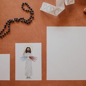 May include: A white card with a watercolor illustration of Jesus with the words "Jesus I trust in you" printed below. There is a rosary, a white ribbon with circles, and a wooden cross with a silver figure of Jesus on it on a brown surface.