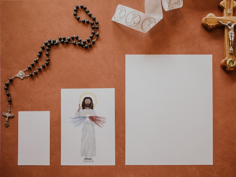 May include: A white card with a watercolor illustration of Jesus with the words "Jesus I trust in you" printed below. There is a rosary, a white ribbon with circles, and a wooden cross with a silver figure of Jesus on it on a brown surface.