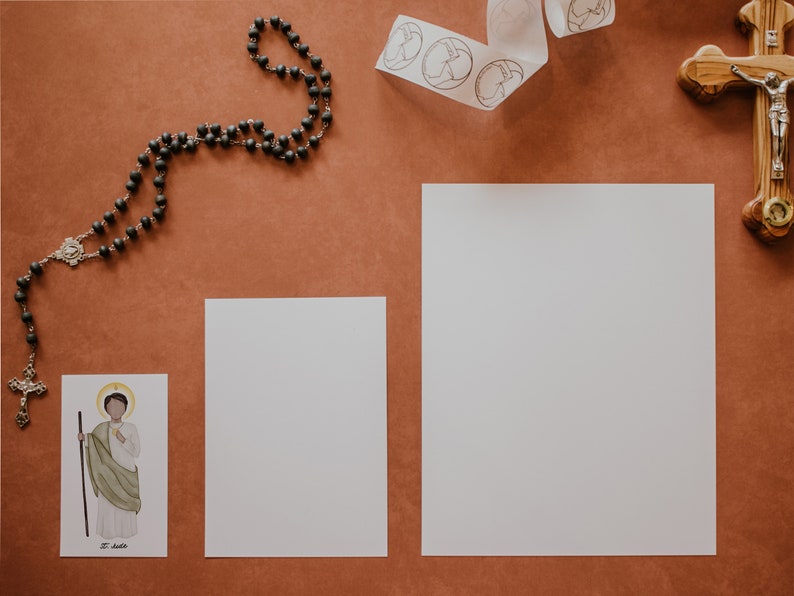 May include: A flat lay of religious items on a brown surface. A black rosary, a white card with a saint's image, two blank white cards, and a wooden cross with a silver figure of Jesus.