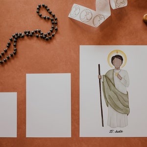 May include: A white card with a watercolor illustration of a saint holding a staff and a golden medallion. The saint is wearing a white robe with a green cloak. The card has the text "St. Jude" at the bottom.