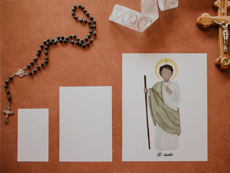 May include: A white card with a watercolor illustration of a saint holding a staff and a golden medallion. The saint is wearing a white robe with a green cloak. The card has the text "St. Jude" at the bottom.
