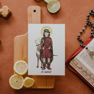 May include: A watercolor illustration of Saint Hubert, a patron saint of hunters, with a stag and a cross. The saint is depicted wearing a green cloak and red tunic, holding a bow and arrow. The illustration is on a white background with the text "St. Hubert" at the bottom.