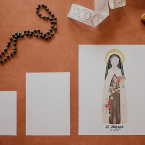 Puede incluir: Una impresi&oacute;n de arte imprimible blanca con una ilustraci&oacute;n de acuarela de Santa Teresa de Lisieux, una santa cat&oacute;lica. La ilustraci&oacute;n la muestra con un h&aacute;bito blanco con una faja marr&oacute;n y un ramo de rosas. El texto "Santa Teresa de Lisieux" est&aacute; impreso debajo de la ilustraci&oacute;n.
