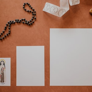 Puede incluir: Una disposici&oacute;n plana de art&iacute;culos religiosos sobre una superficie marr&oacute;n. Hay un rosario, una tarjeta peque&ntilde;a con una imagen de un santo, dos tarjetas blancas en blanco y una cruz de madera con una figura de Jes&uacute;s de plata.