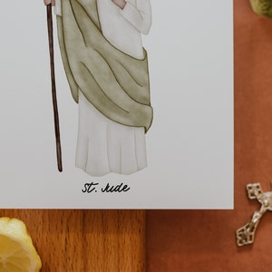 May include: A watercolor illustration of a saint holding a staff. The saint is wearing a white robe with a green cloak. The illustration is on a white background with the text "St. Jude" written below.