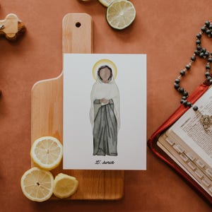 May include: A watercolor illustration of St. Junia, depicted in a white head covering and gray robe, with a gold halo. The artwork is on a white card, resting on a wooden cutting board with lemon slices. A rosary and open book are also visible.