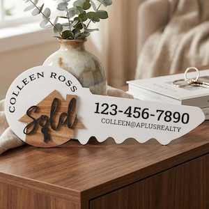 May include: A decorative wooden key-shaped sign with the words "Colleen Ross" and a phone number. The sign features a wooden house and the word "Sold". A vase with greenery sits behind the sign.