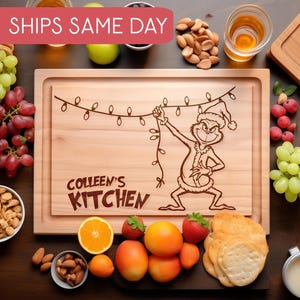 May include: Wooden chopping board featuring a Grinch design holding Christmas lights. The words "COLLEEN'S KITCHEN" are engraved below. Grapes, nuts, fruit, and crackers surround the board, creating a festive display.