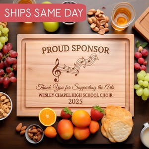 Puede incluir: Una tabla de cortar de madera con el texto "PROUD SPONSOR" y "Thank You for Supporting the Arts" está rodeada de uvas, nueces y frutas. La tabla también presenta un diseño de notas musicales y el texto "WESLEY CHAPEL HIGH SCHOOL CHOIR 2025".