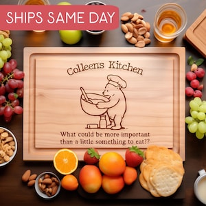 以下が含まれることがあります： 「Colleen's Kitchen」の文字とクマのシェフのイラストが描かれた木製のまな板。ブドウ、ナッツ、クラッカー、フルーツに囲まれています。「SHIPS SAME DAY」の文字が上部にあります。