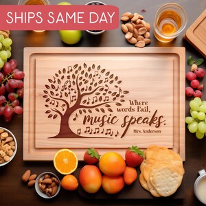 May include: A wooden cutting board with a tree design and the quote "Where words fail, music speaks."  Mrs. Anderson is engraved below the quote. The board is surrounded by various food items, including grapes, nuts, and fruit.