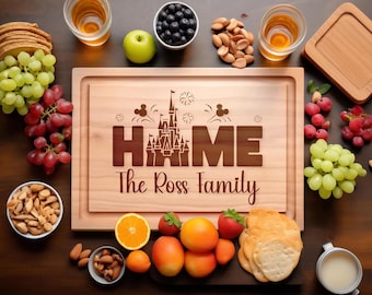 Puede incluir: Una tabla de cortar rectangular de madera con un diseño grabado a láser de la silueta de un castillo que sustituye a la letra O en la palabra HOME, con el texto The Ross Family debajo. Rodeada de uvas, frutos secos, fruta y galletas.