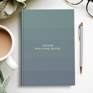 May include: A journal with a gradient blue-green cover, the text "colleen's brain dump journal" is centered. A silver pen, a cup of coffee, and eyeglasses are next to the journal. The journal is a notebook.