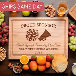 Puede incluir: Tabla de cortar de madera con el texto "PROUD SPONSOR" y "Thank You for Supporting Our Team". Presenta un diseño de pompón y megáfono. También se ve el texto "WESLEY CHAPEL HIGH SCHOOL CHEER 2025". La imagen incluye uvas, nueces y fruta.