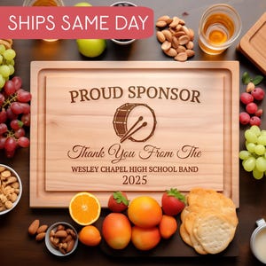 Puede incluir: Tabla de cortar de madera con la inscripción "PROUD SPONSOR" y "Thank You From The WESLEY CHAPEL HIGH SCHOOL BAND 2025". Rodeada de aperitivos y frutas, como uvas, almendras y galletas saladas. Ideal para eventos.