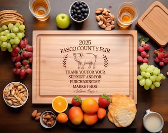 May include: A rectangular wooden cutting board engraved with the text 2025 PASCO COUNTY FAIR, an illustration of a pig, and the words THANK YOU FOR YOUR SUPPORT AND FOR PURCHASING MY MARKET HOG, signed by Colleen Ross, surrounded by assorted fruit.