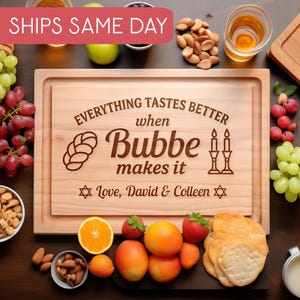 May include: A wooden cutting board with a carved message that reads "Everything tastes better when Bubbe makes it." The board is surrounded by food items like grapes, almonds, and fruit. The text "Love, David & Colleen" is also carved on the board.