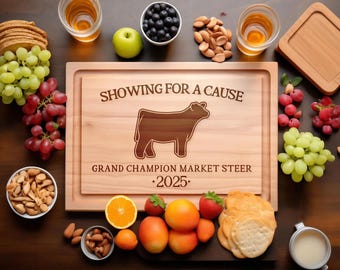 May include: A rectangular wooden cutting board engraved with the text SHOWING FOR A CAUSE, GRAND CHAMPION MARKET STEER, and 2025, featuring a steer silhouette. It is surrounded by grapes, apples, blueberries, nuts, oranges, and crackers.