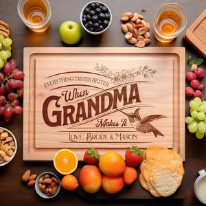 Puede incluir: Una tabla de cortar rectangular de madera grabada con el texto "Everything Tastes Better When Grandma Makes It" y "Love, Brody & Mason" junto a un diseño de colibrí y flores, rodeada de uvas, nueces, fruta y galletas.