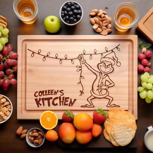 May include: A rectangular wooden chopping board featuring a laser-engraved illustration of a green creature wearing a Santa hat and holding a string of Christmas lights, with the text "COLLEEN'S KITCHEN" engraved in the lower left corner.
