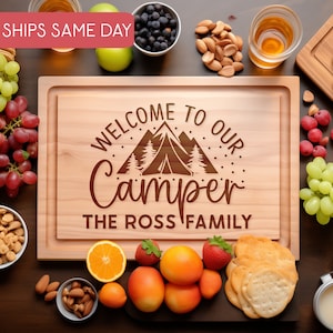 May include: A wooden cutting board with the text "Welcome to our Camper The Ross Family" engraved on it. The board is surrounded by various food items, including grapes, blueberries, almonds, crackers, and fruit.