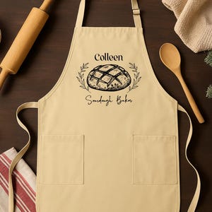May include: A beige apron with two front pockets, featuring the name "Colleen" above a bread illustration and the words "Sourdough Baker." A rolling pin, wooden spoon, and ingredients are visible in the background.
