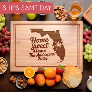 May include: A personalized wooden cutting board with the text "Home Sweet Home The Andersons 2024" and an outline of the state of Florida. The board is surrounded by various food items, including grapes, almonds, crackers, and fruit.