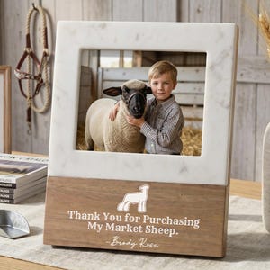 May include: A rectangular photo frame with a white marble top and a wooden base. The frame holds a photo of a boy with a sheep. The text on the base reads "Thank You for Purchasing My Market Sheep."