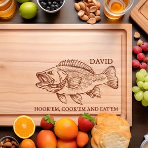 May include: A rectangular light-coloured wooden cutting board featuring a laser-engraved illustration of a fish in the centre. The name DAVID is engraved above the fish, and the text HOOK'EM, COOK'EM AND EAT'EM is engraved below the fish.