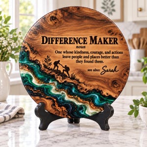 May include: A round wooden plaque on a black stand featuring a natural wood grain, a swirling green and white resin river design, and a silhouette of two hikers. The text reads: Difference Maker, noun, One whose kindness, courage, and actions leave people and places better than they found them, see also: Sarah.