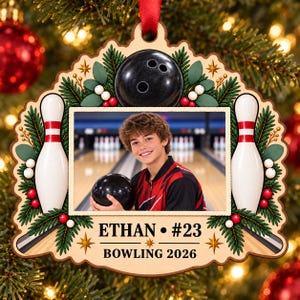 May include: A wooden Christmas ornament featuring a central photo of a person holding a black bowling ball at a bowling alley. The frame is decorated with bowling pins, pine branches, red and white berries, and the text ETHAN #23 BOWLING 2026.