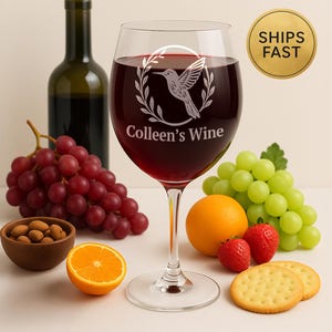 Puede incluir: Una copa de vino llena de vino tinto, con un diseño de colibrí y el texto "Colleen's Wine". La imagen incluye una botella de vino, uvas, rodajas de naranja, fresas y galletas, lo que sugiere un maridaje de vino y comida.