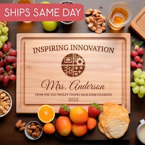 Puede incluir: Una tabla de cortar de madera con el texto "INSPIRING INNOVATION" y "Mrs. Anderson" grabado. La tabla está rodeada de fruta, frutos secos y galletas saladas. También está grabado el texto "FROM THE 2025 WESLEY CHAPEL HIGH STEM STUDENTS" y "2025".