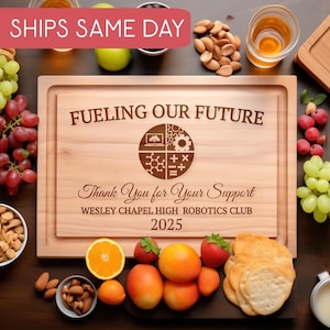 Puede incluir: Una tabla de cortar de madera con un borde empotrado, grabada con el texto "FUELING OUR FUTURE" y "Thank You for Your Support". La tabla está rodeada de uvas, nueces, frutas y galletas saladas. También está grabado el texto "WESLEY CHAPEL HIGH ROBOTICS CLUB 2025".