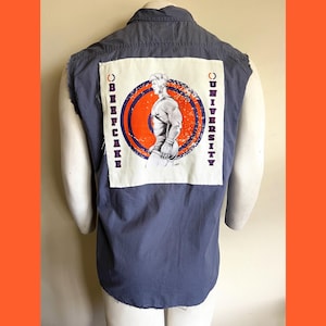 May include: A sleeveless blue denim shirt with a white patch on the back. The patch features a muscular figure in an orange circle with the text "BEERCAKE UNIVERSITY" on either side.