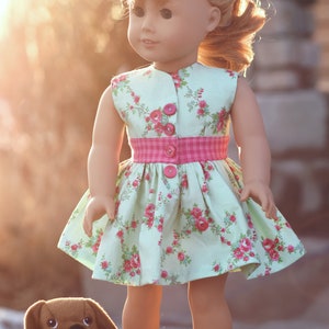 Pode incluir: Uma boneca vestindo um vestido verde claro com estampa floral rosa e uma faixa rosa. A boneca tem cabelo loiro e olhos azuis. A boneca está segurando um cachorro de pelúcia marrom.