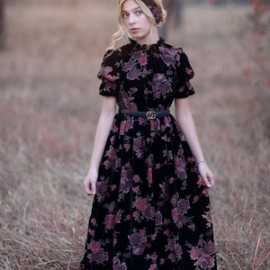 Puede incluir: Vestido de terciopelo negro con estampado floral en tonos rojos y morados. El vestido presenta mangas cortas, cuello alto y cinturón negro. Una diadema floral a juego completa el conjunto.