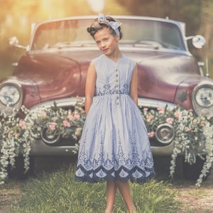 Pode incluir: Uma menina jovem com um vestido azul e branco está em frente a um carro antigo decorado com flores. O carro é de cor vinho e tem um pára-choques branco. A menina está sorrindo e olhando para a câmera.