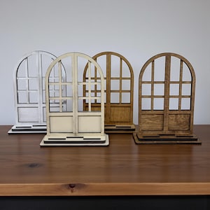 Peut inclure: Quatre décorations de fenêtres en bois en forme d'arche, dans des tons de blanc et de marron. Chacune a une base rectangulaire et un motif de fenêtre en grille. Les fenêtres sont exposées sur une surface en bois foncé.